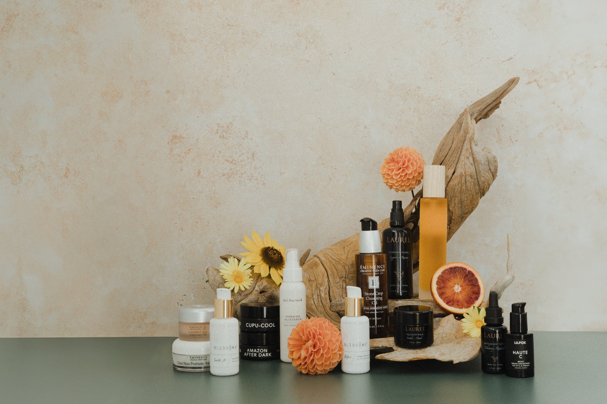 Collection of organic skincare products fro Eminence organics, Laurel, Lilfox, Blissoma, etc. on a table with decorative elements against a beige wall.