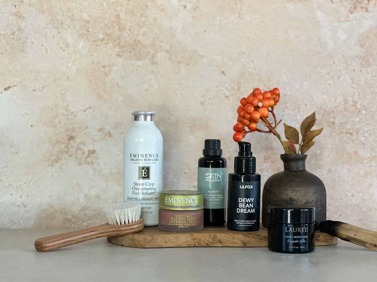 A curated collection of skin care products arranged on a round wooden tray against a textured beige stone wall. The products include exfoliants from various clean beauty brands, alongside a wooden hairbrush and a small vase with orange berries.