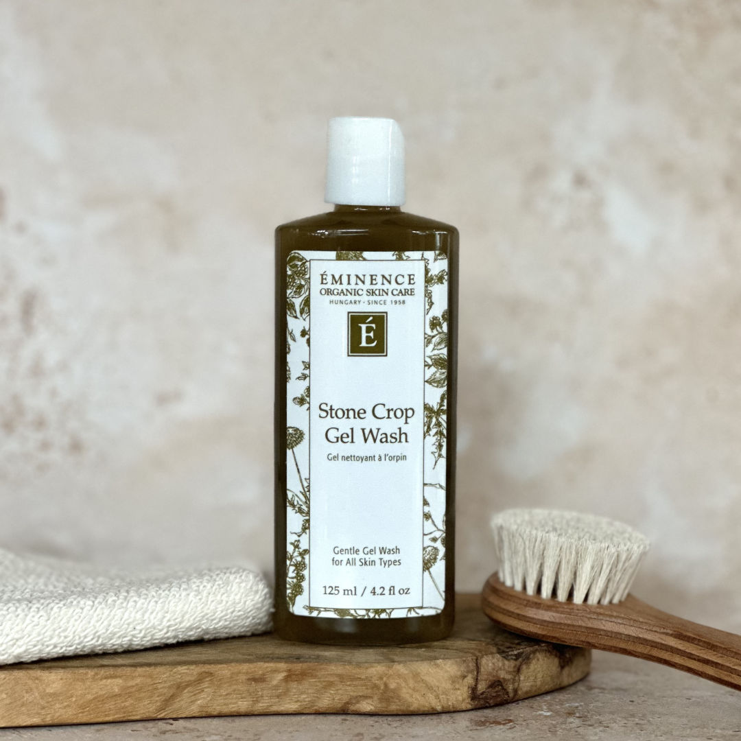 Bottle of Eminence Stone Crop Gel Wash on a wooden surface with a brush and towel in the background.